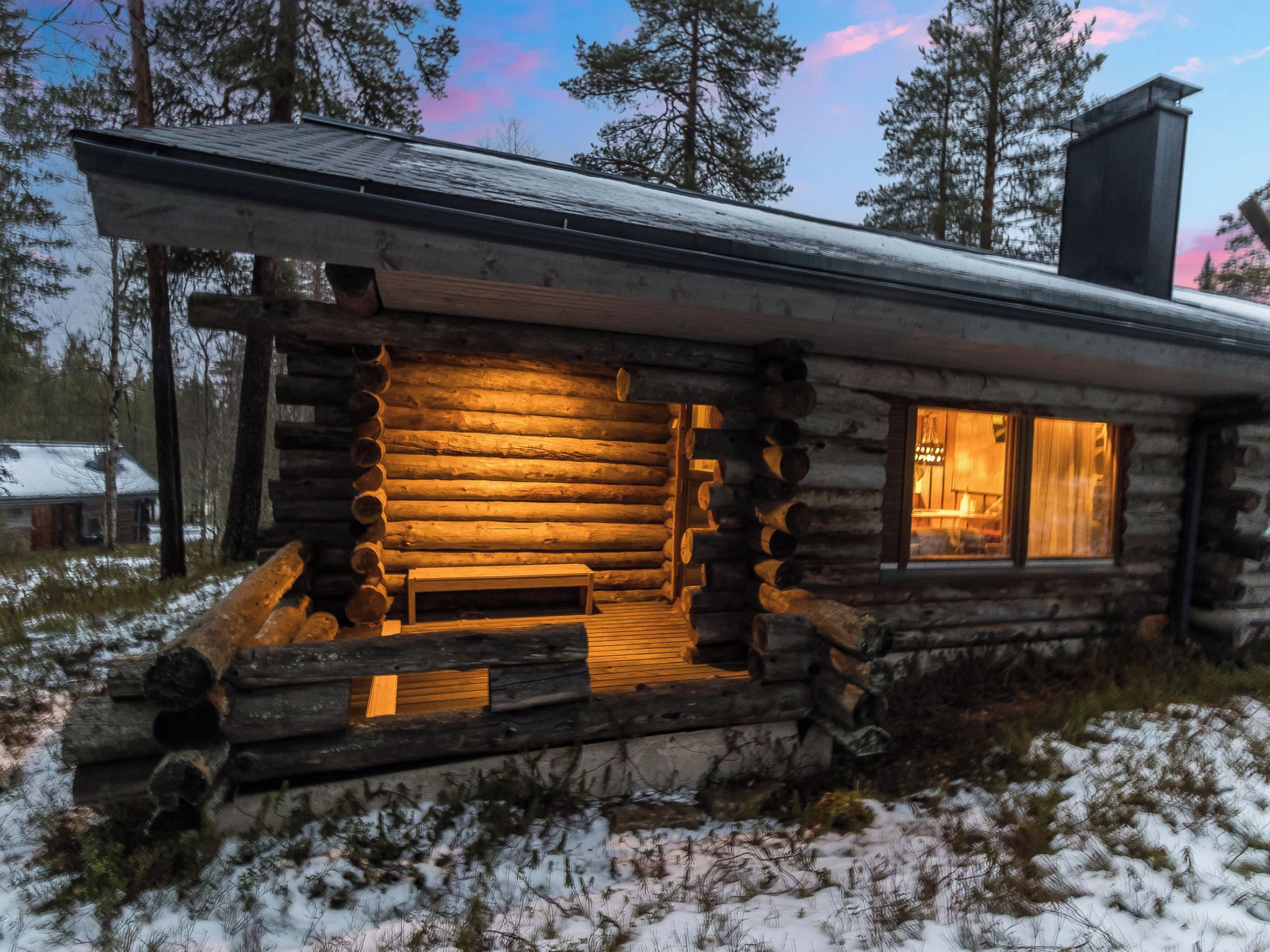 Hirsimökki, jonka ulkoseinät ovat puuta, siinä on kuisti penkkeineen ja ikkunassa verhot.