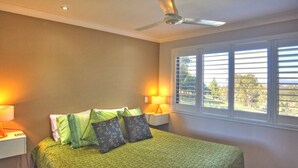 A bedroom with a bed, two bedside lamps, a ceiling fan, and a window with blinds.