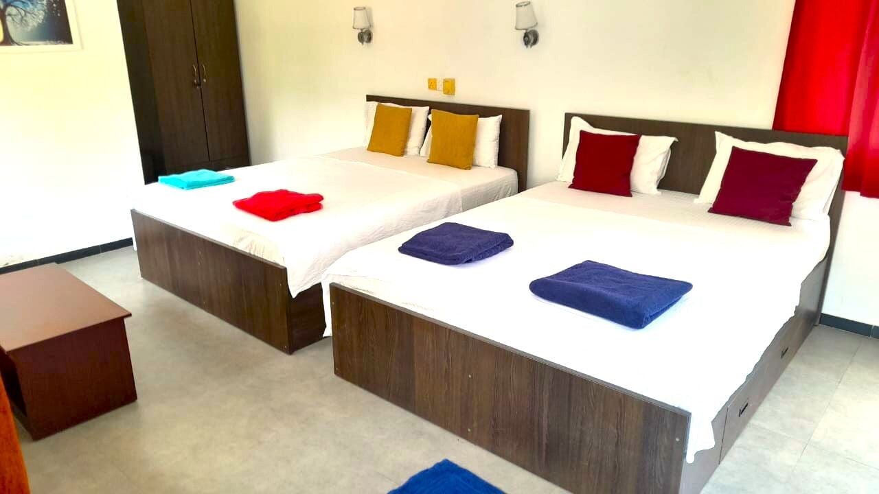 A hotel room with two beds, each with a wooden headboard, white bedding, and colorful pillows. There is a small wooden nightstand and a red curtain on the right side.