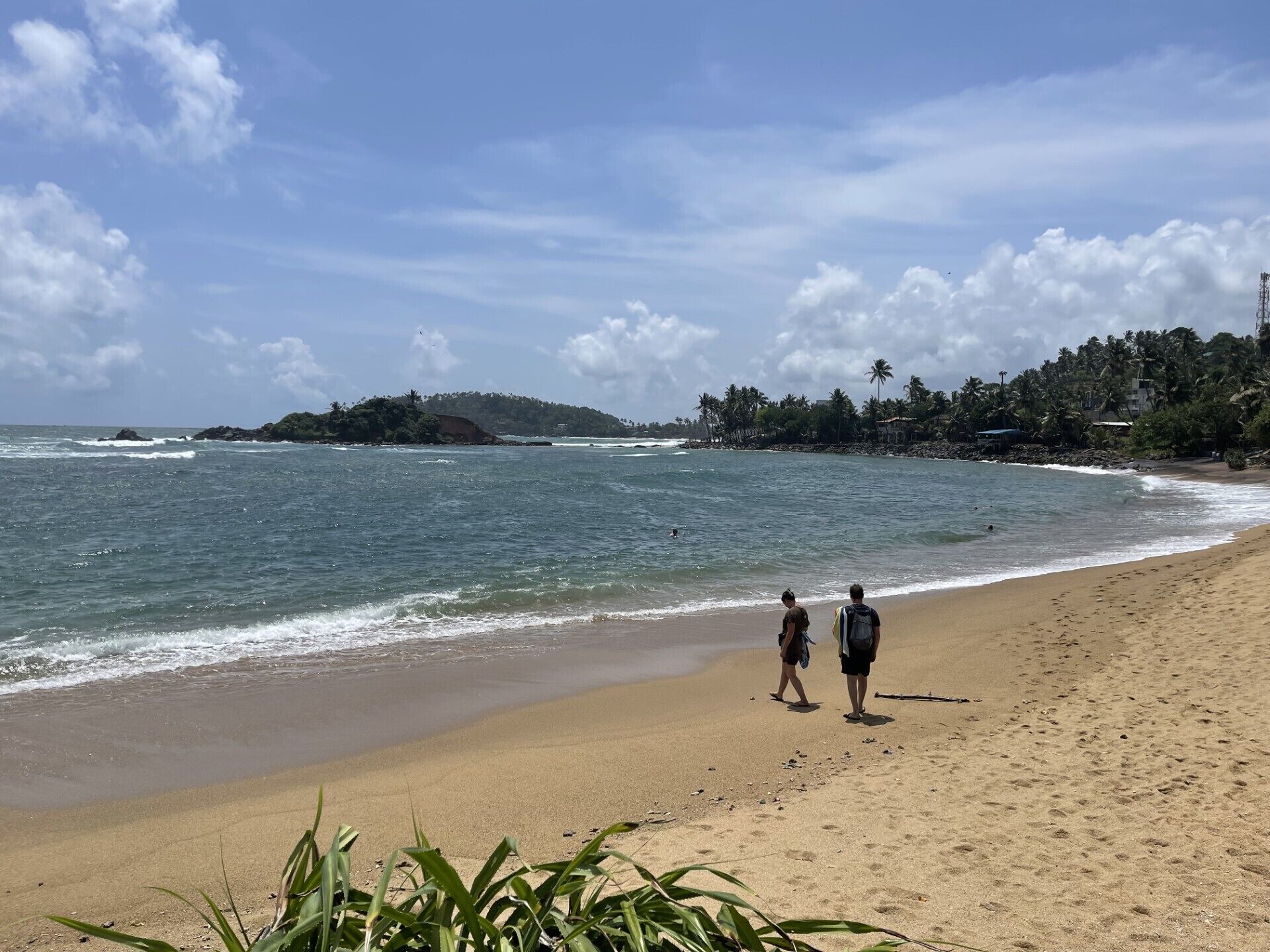 A beach with two people walking along the shore, a sandy beach, and a distant island with vegetation.