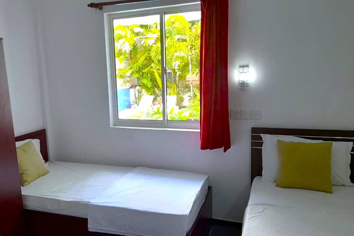 A small, clean room with a single bed, a window with red curtains, and a view of greenery outside.