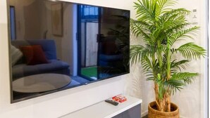 Un televisor de pantalla plana montado en una pared blanca, una planta en maceta a su lado y un control remoto sobre el mueble de la TV.
