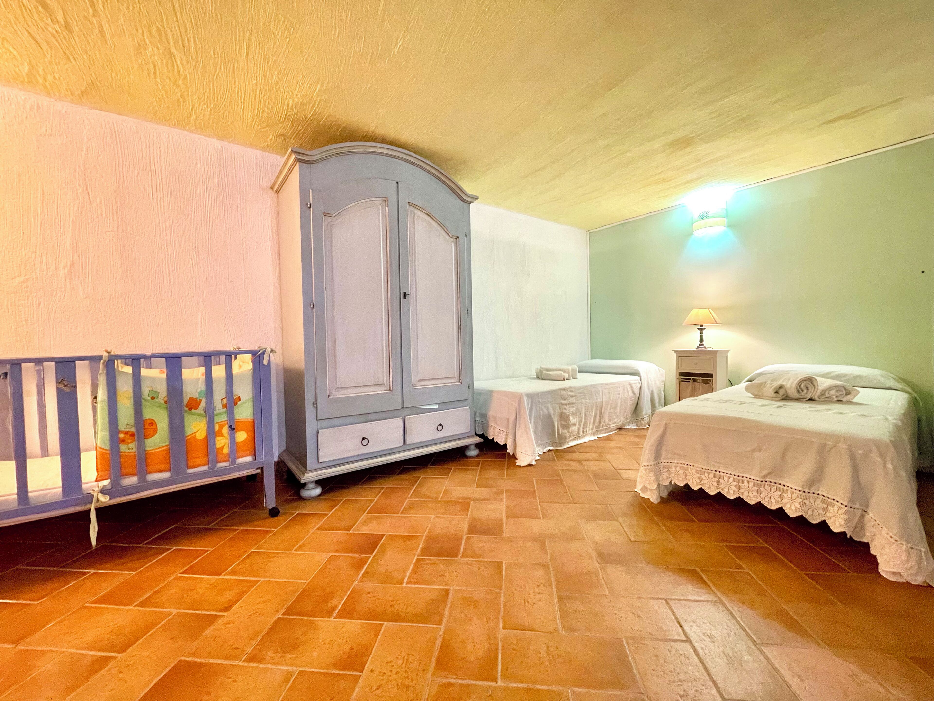 A bedroom with a crib, two beds, a wardrobe, and a lamp.