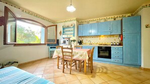 A compact kitchen with blue cabinets, a dining table with chairs, a window, and a tiled floor. - " Villa Teresa con patio " (Bari Sardo)