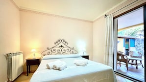A neatly made bed with white linens and towels, a radiator, a nightstand with a lamp, and a view of an outdoor patio area. - " Villa Teresa con patio " (Bari Sardo)