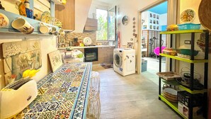 A compact kitchen with a washing machine, a dining table with a colorful tiled runner, and various kitchen utensils and dishes on shelves. - "  La  Corte   Azzurra  " (Bari Sardo)