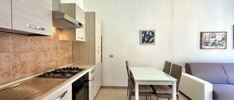 Una cocina moderna con estufa a gas, horno y armarios de colores claros. Hay una mesa de comedor con sillas y un sofá en la zona de estar.