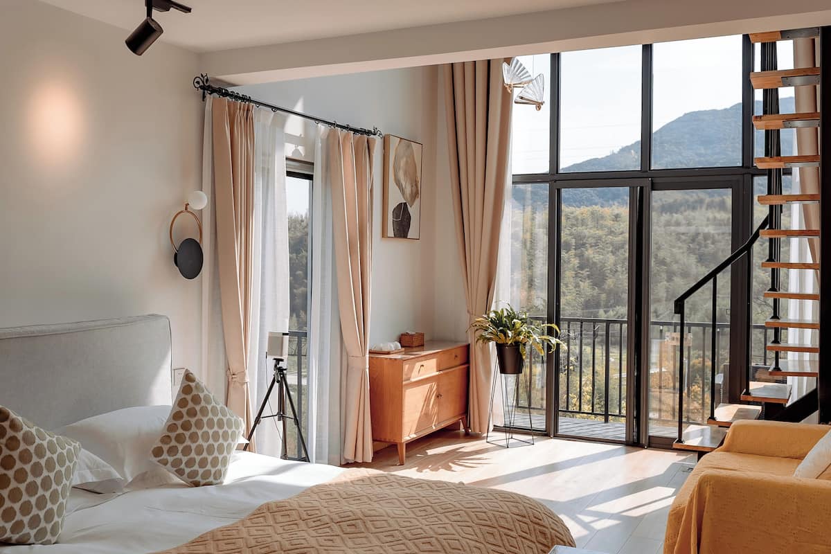 A bedroom with a large window, a bed, a dresser, a plant, and a staircase.