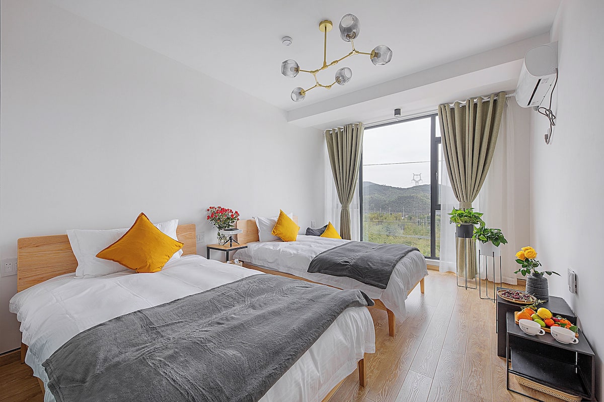 A hotel room with two beds, a modern chandelier, a large window with curtains, a small table with a vase of flowers, and a view of the outdoors.