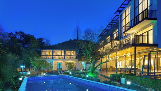 A modern hotel with a swimming pool, outdoor seating, and a well-lit entrance.