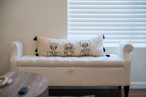 A white sofa with a pillow featuring cartoon faces, positioned in front of a window with blinds.