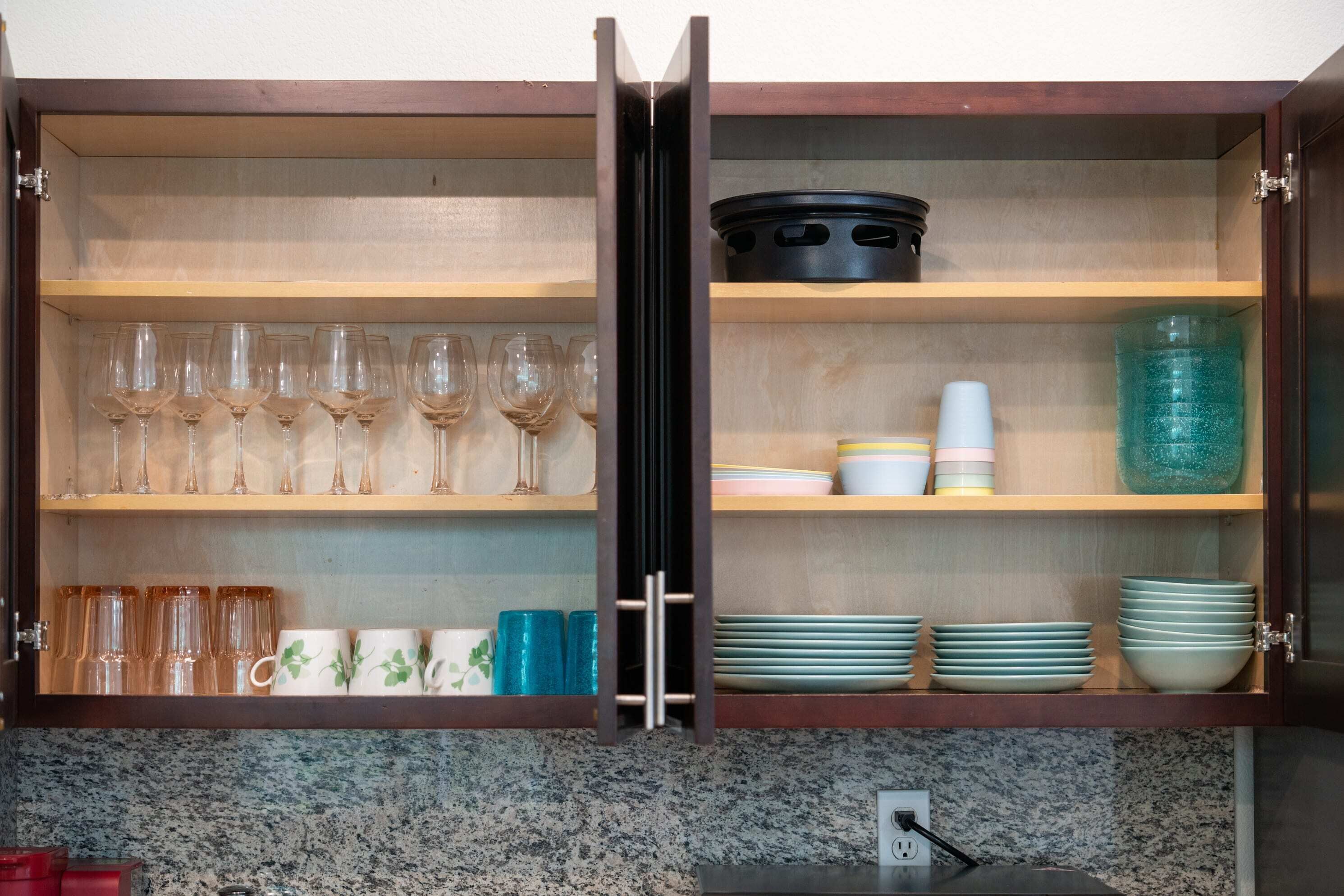 A kitchen cabinet with shelves holding wine glasses, cups, and plates.