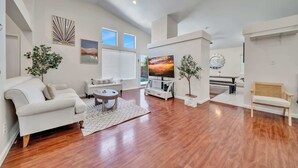 A spacious living area with a white sofa, a coffee table, a TV, and a dining table with chairs. There are two potted plants and a wall clock. - @marbella Lane - 4BR Lavish Castle Retreat (Indio)