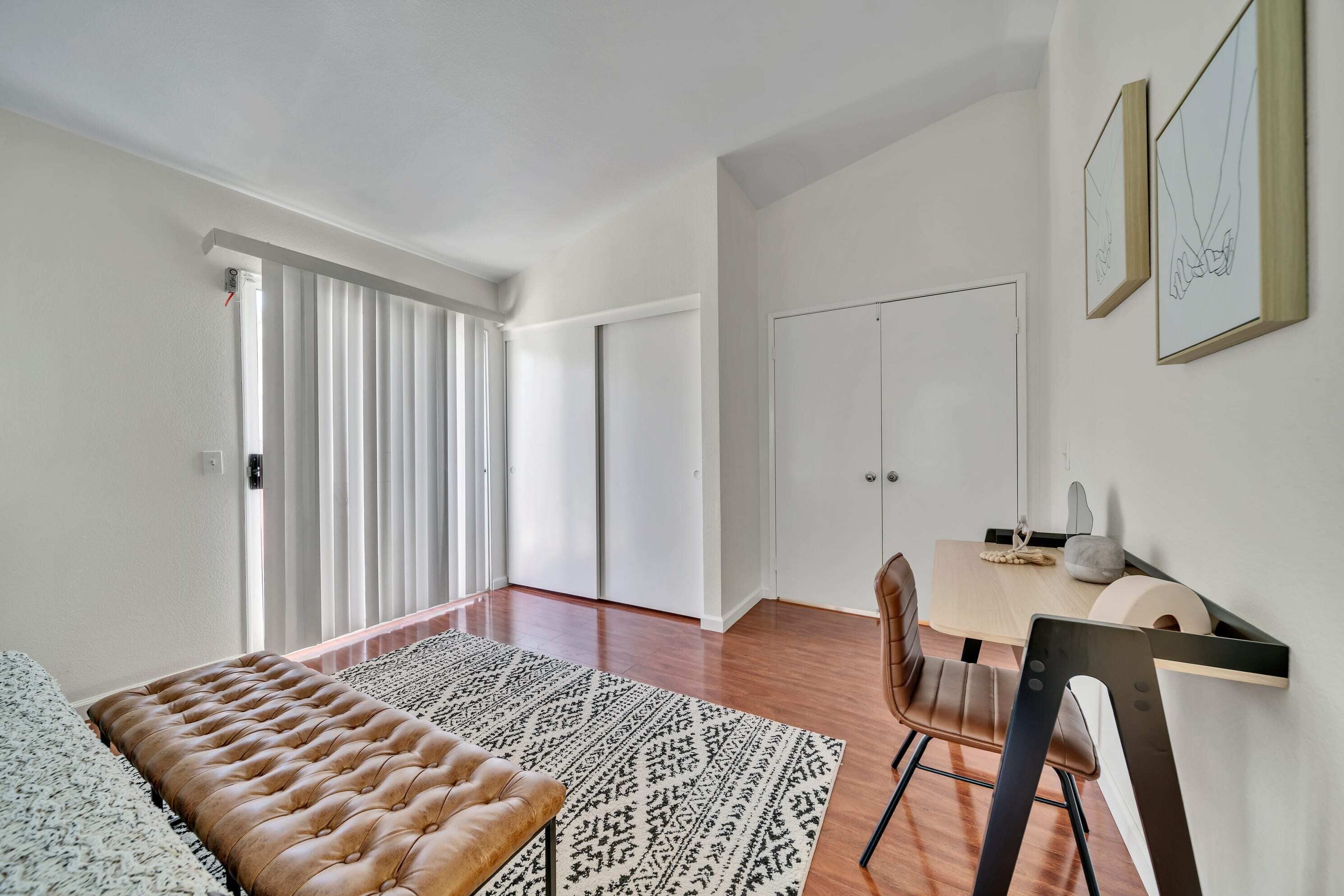 A modern bedroom with a tufted headboard, a patterned rug, a wooden floor, a desk with a chair, and a sliding door.