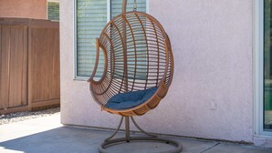A wicker egg chair with a cushion, mounted on a circular base, positioned on a concrete patio against a building with a wooden fence and a window with blinds. - @marbella Lane - 4BR Lavish Castle Retreat (Indio)