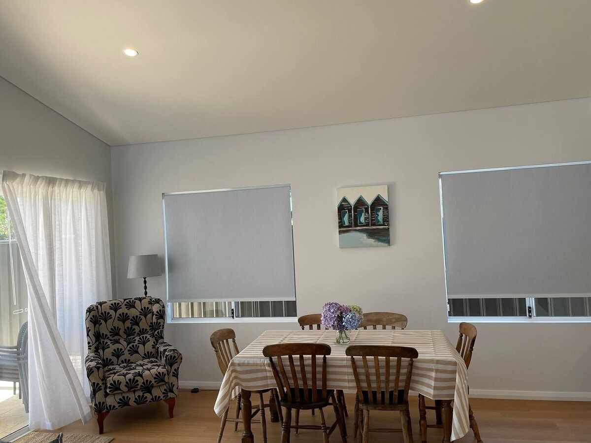 Un coin repas avec une table dressée pour six personnes, entourée de six chaises en bois. Un fauteuil unique, orné d’un motif floral, se trouve à gauche. La pièce est dotée de deux fenêtres avec des stores, d’une lampe fixée au mur et d’un petit objet décoratif placé au-dessus de la table.