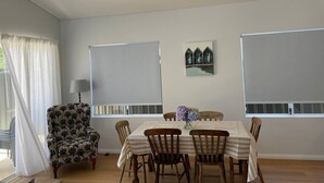 A dining area with a table set for six, surrounded by six wooden chairs. There is a single armchair with a floral pattern to the left. The room has two windows with blinds, a wall-mounted lamp, and a small decorative piece above the table. - The Front House (Augusta)