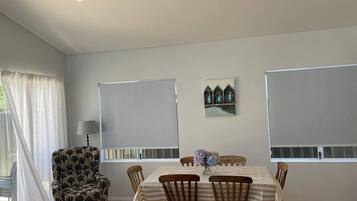 Un coin repas avec une table dressée pour six personnes, entourée de six chaises en bois. Un fauteuil unique, orné d’un motif floral, se trouve à gauche. La pièce est dotée de deux fenêtres avec des stores, d’une lampe fixée au mur et d’un petit objet décoratif placé au-dessus de la table.