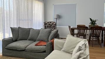 Un salon moderne avec un canapé gris, un canapé blanc, une table à manger avec des chaises et une grande fenêtre ornée de rideaux légers.