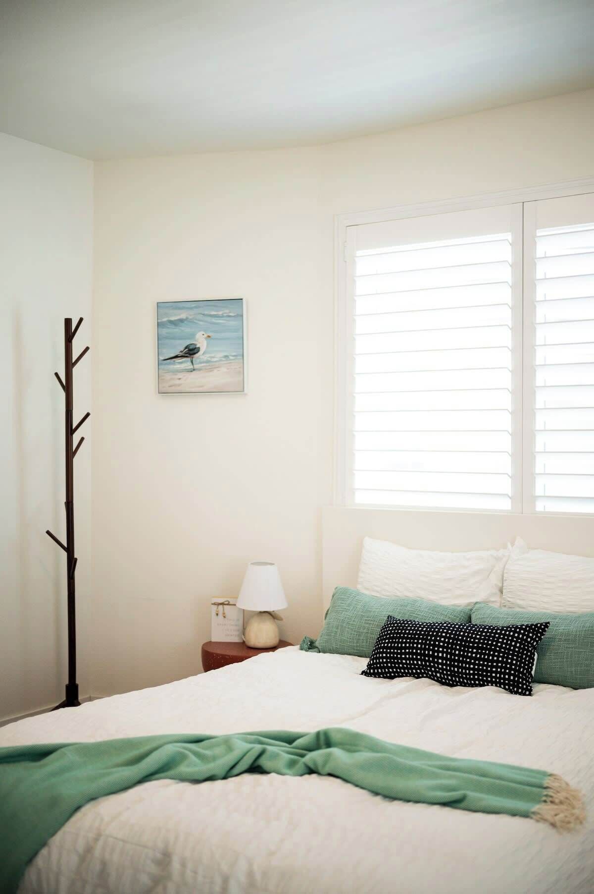 A bedroom with a bed, a nightstand, a lamp, a window with blinds, and a wall-mounted picture.