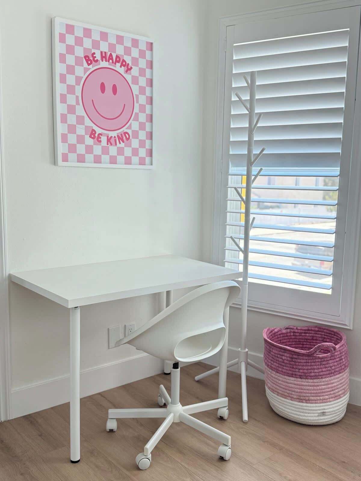 A white desk with a white chair, a pink and white checkered wall art with a smiley face and the words "BE HAPPY BE KIND", and a white window with blinds.