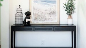 A black console table with a framed landscape painting, a black dog with a 'WELCOME' sign, a birdcage, and a hanging light fixture. - Cozy Retreat Near Downtown Perfect for Families (Long Beach)