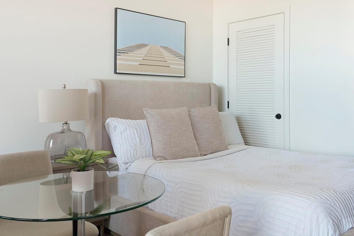 Une chambre d’hôtel bien rangée, avec un lit, une table en verre ornée d’une plante, une lampe et un tableau encadré accroché au mur.