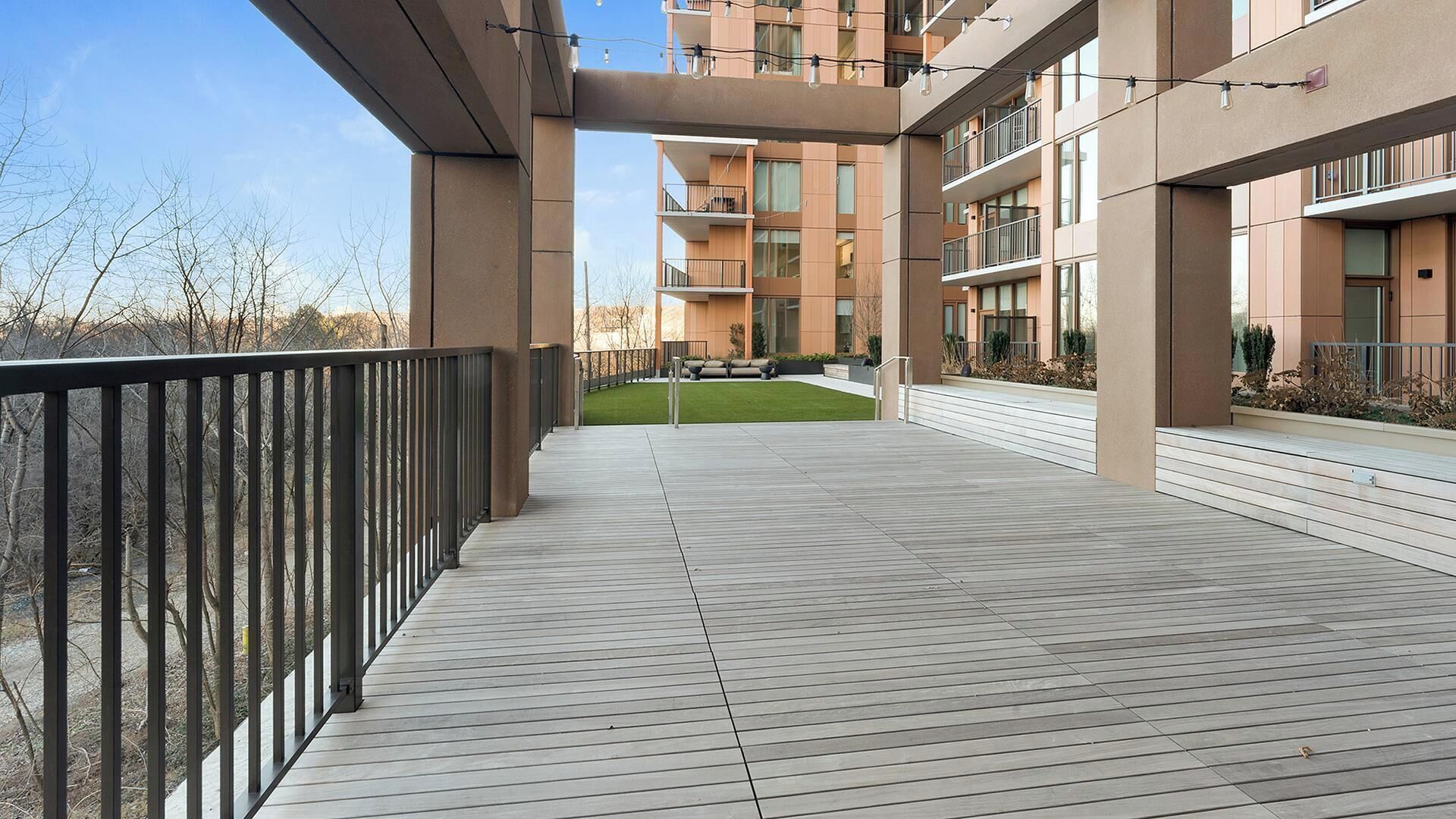A modern outdoor balcony with a wooden deck, metal railings, and a view of a multi-story building with balconies.