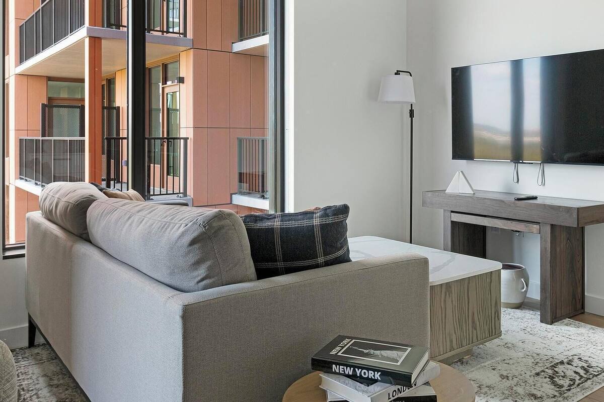 Un salon moderne comprenant un canapé, un téléviseur à écran plat, une petite table d’appoint en bois et une fenêtre donnant sur un immeuble.