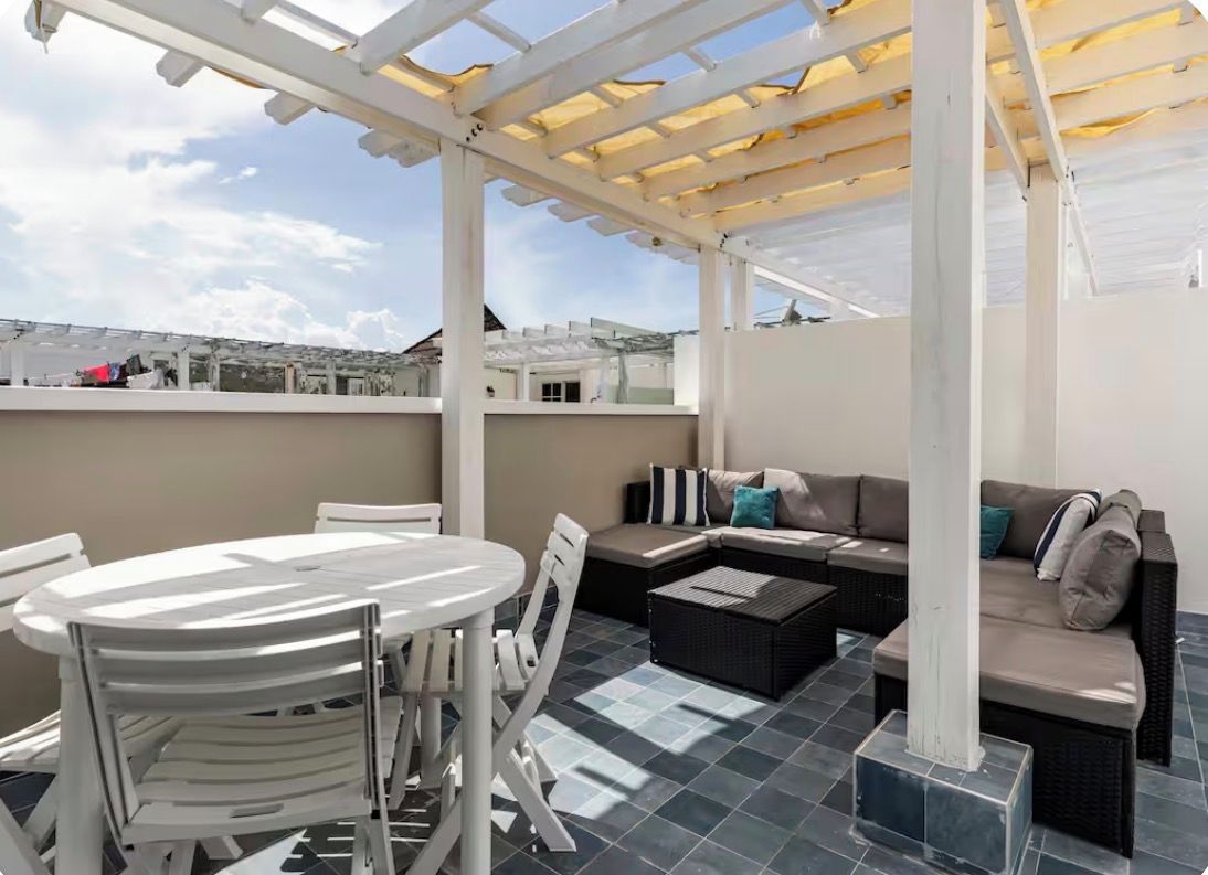 An outdoor patio area with a round table, white chairs, a sofa, and a pergola structure.