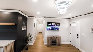 A modern hallway with a wooden floor, a black accent wall, a mounted TV displaying a channel guide, and a ceiling light fixture.
