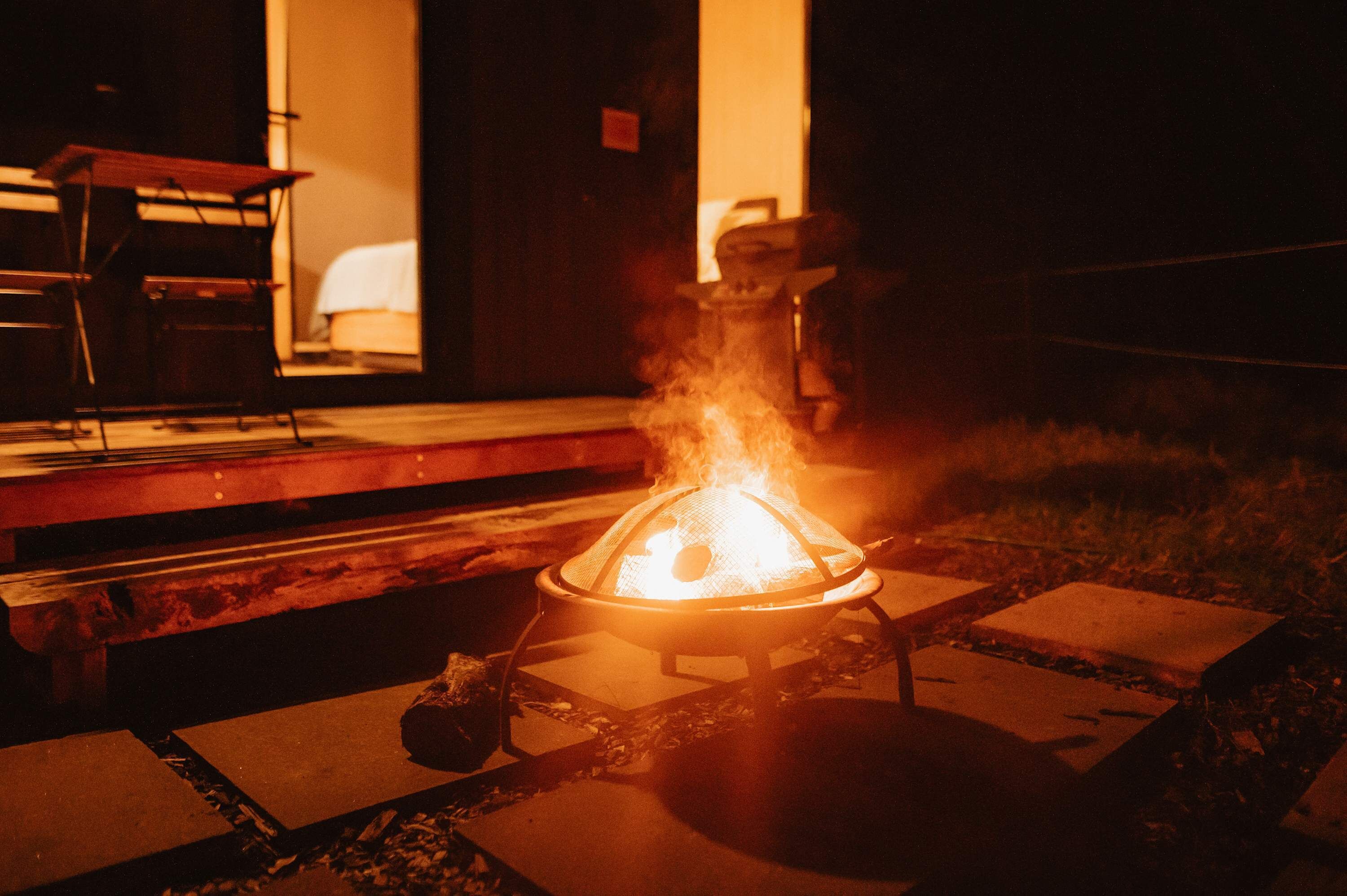En lägereld med ett metallgaller, omgiven av betongplattor och en bänk, i en svagt upplyst utomhusmiljö.