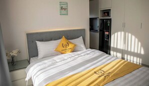 A hotel room with a bed, a nightstand, a vase of flowers, a refrigerator, and a wall with a sign. - Grand Park Hotel & Apartment Phu My Hung (Ho Chi Minh City)