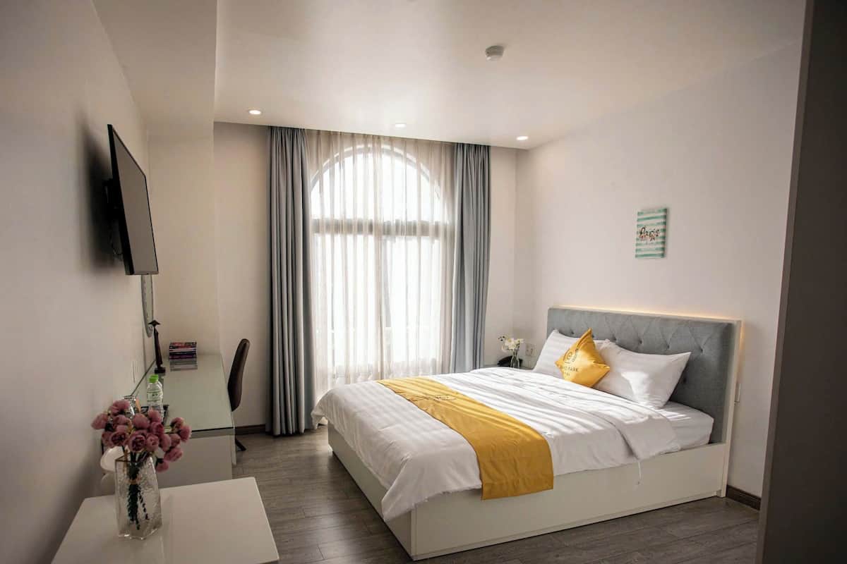 Quarto de hotel com cama, escrivaninha, cadeira, televisão na parede e janela com cortinas.