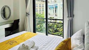 A hotel room with a bed, a desk, a chair, a large window with curtains, and a view of buildings outside.