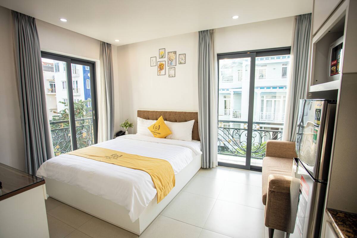 Quarto de hotel com uma cama grande, um pequeno sofá, um frigorífico e uma varanda com vista para edifícios.