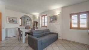 A spacious living area with a grey sofa, a dining table, and chairs. There are two windows with wooden frames and a wall-mounted light fixture. - IL BORGHETTO - VILLINO IL BORGHETTO (Campo nell'Elba)