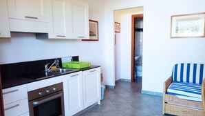 A kitchen with white cabinets, a black countertop, and a green dish rack. There is a wicker chair with a striped cushion in the foreground. - Appartamenti Arcipelago - Appartamenti Arcipelago - Bilocale (30) (Rio dell'Elba)