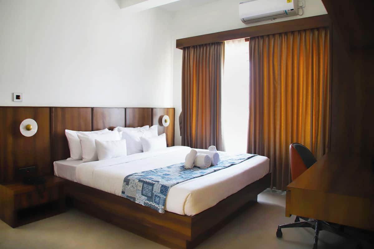 A hotel room with a large bed, wooden headboard, two bedside tables, a desk, and an air conditioning unit.
