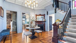 A grand foyer with a wooden floor, a staircase with a dark balustrade, a chandelier, and a fireplace with a mirror above it. - The Royce on Pelham - Luxury Family Estate (Newport)
