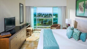 A hotel room with a bed, a TV, a desk, and a chair. There is a large window with a view of the ocean and palm trees.