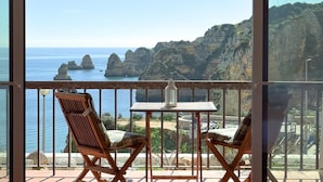 A balcony with two wooden chairs and a small table, overlooking a coastal landscape with rocky cliffs and the sea.