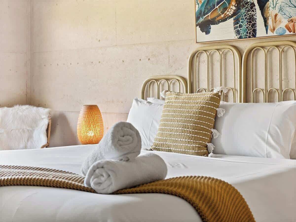 Een net opgemaakt bed met witte lakens, een gerold handdoekje, een textuurrijk beige kussen en een geweven deken.