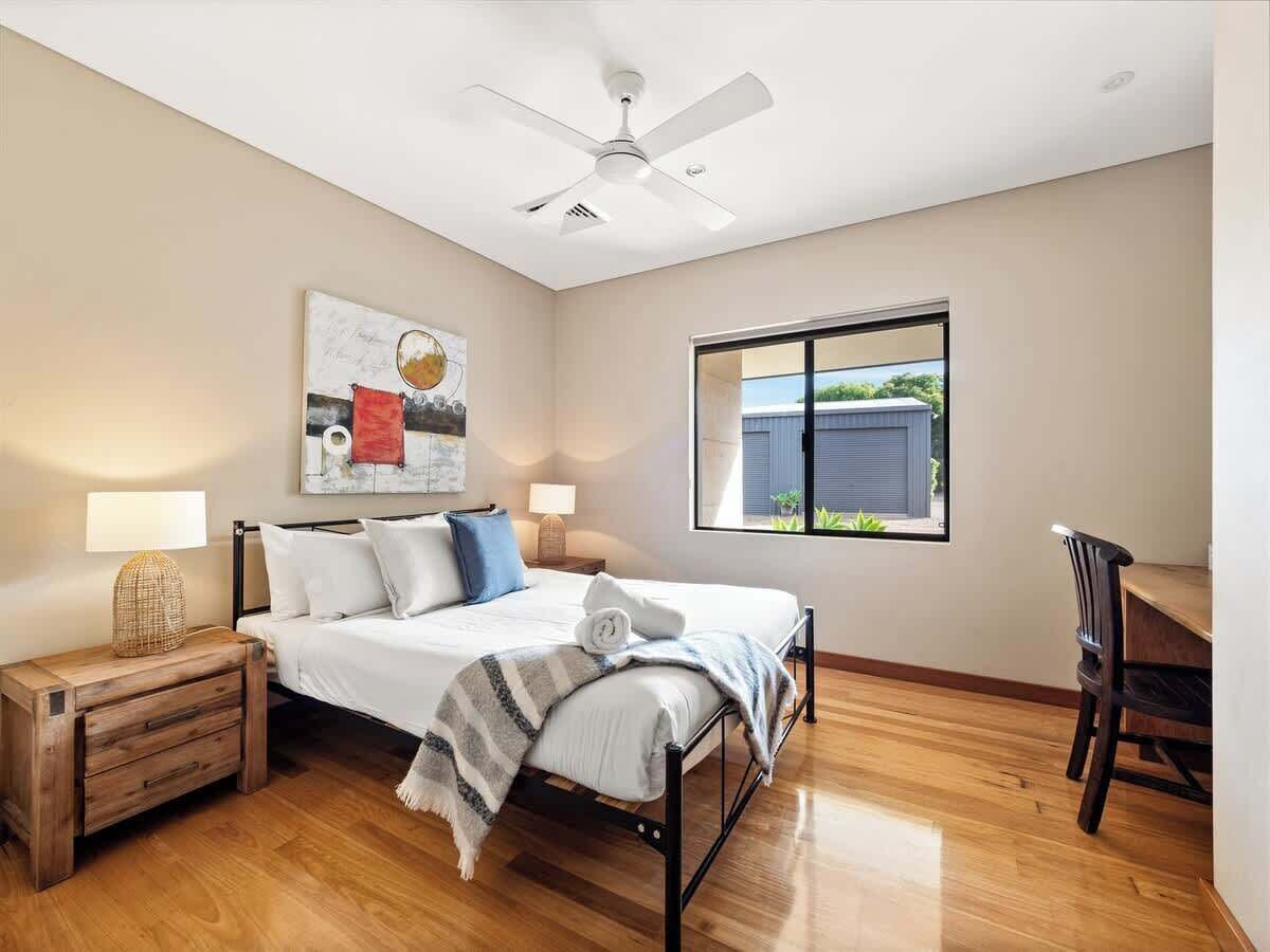 A bedroom with a bed, bedside tables, a desk, a chair, a ceiling fan, a window with a view, and a wall art piece.