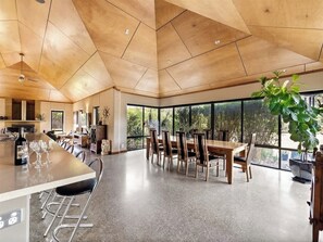 A spacious kitchen with a large island, dining area with chairs, and a view through large windows. - La Dimora—Tranquil Retreat Where Bush Meets Beach (Burnside)