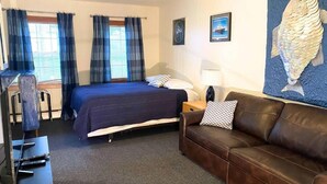 A hotel room with a bed, a brown leather sofa, a desk with a chair, and a ceiling fan. - The Tides Inn (Seward)