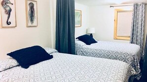 A room with two beds, each with patterned bedding and a dark pillow. There are framed pictures on the wall, a window with blinds, and a curtain. - The Tides Inn (Seward)