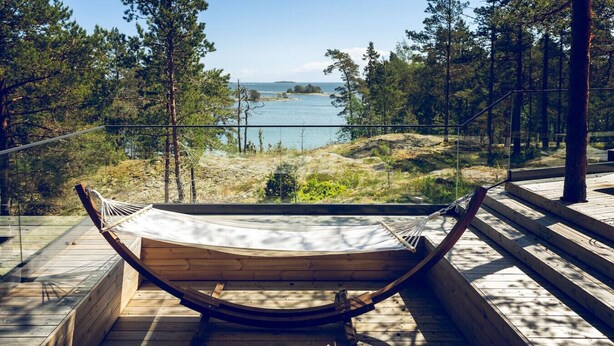 En hängmatta på ett trädäck med utsikt över en sjö, omgiven av träd och en klar himmel.