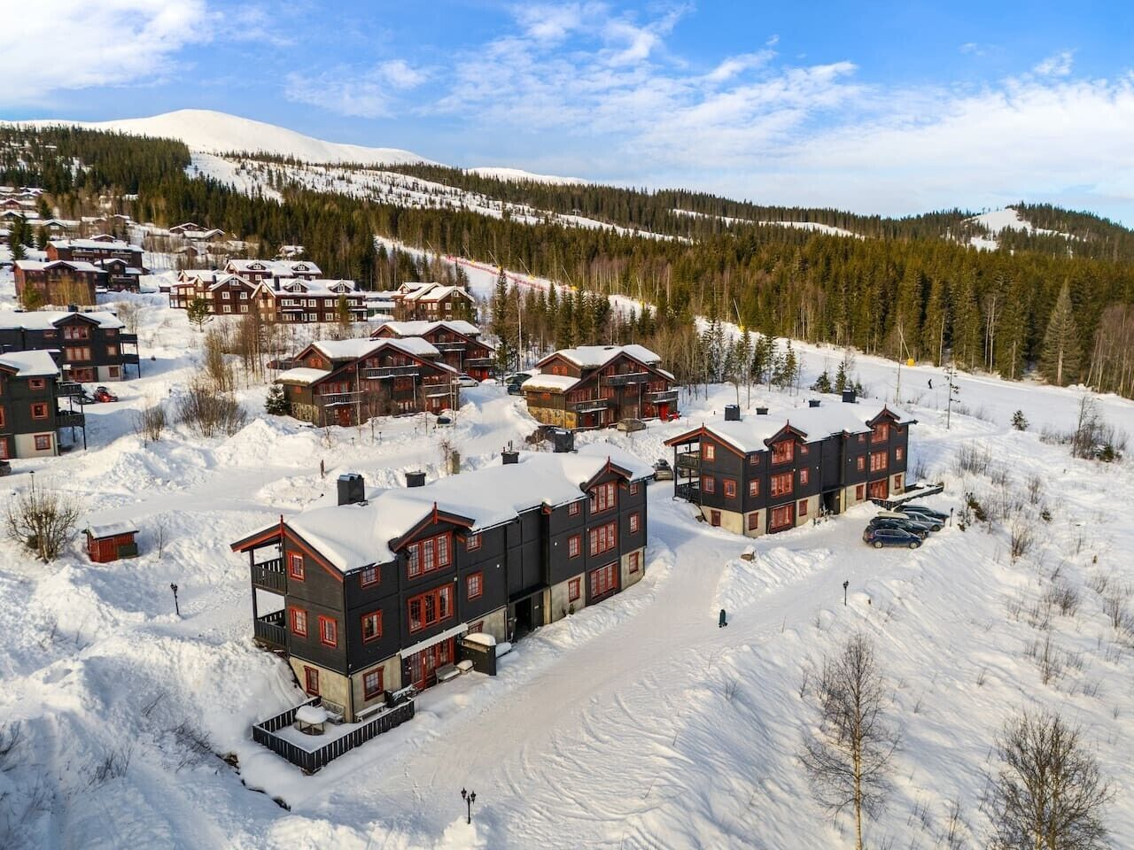 A snow-covered resort with multiple chalet-style buildings, a ski slope in the background, and a clear sky.