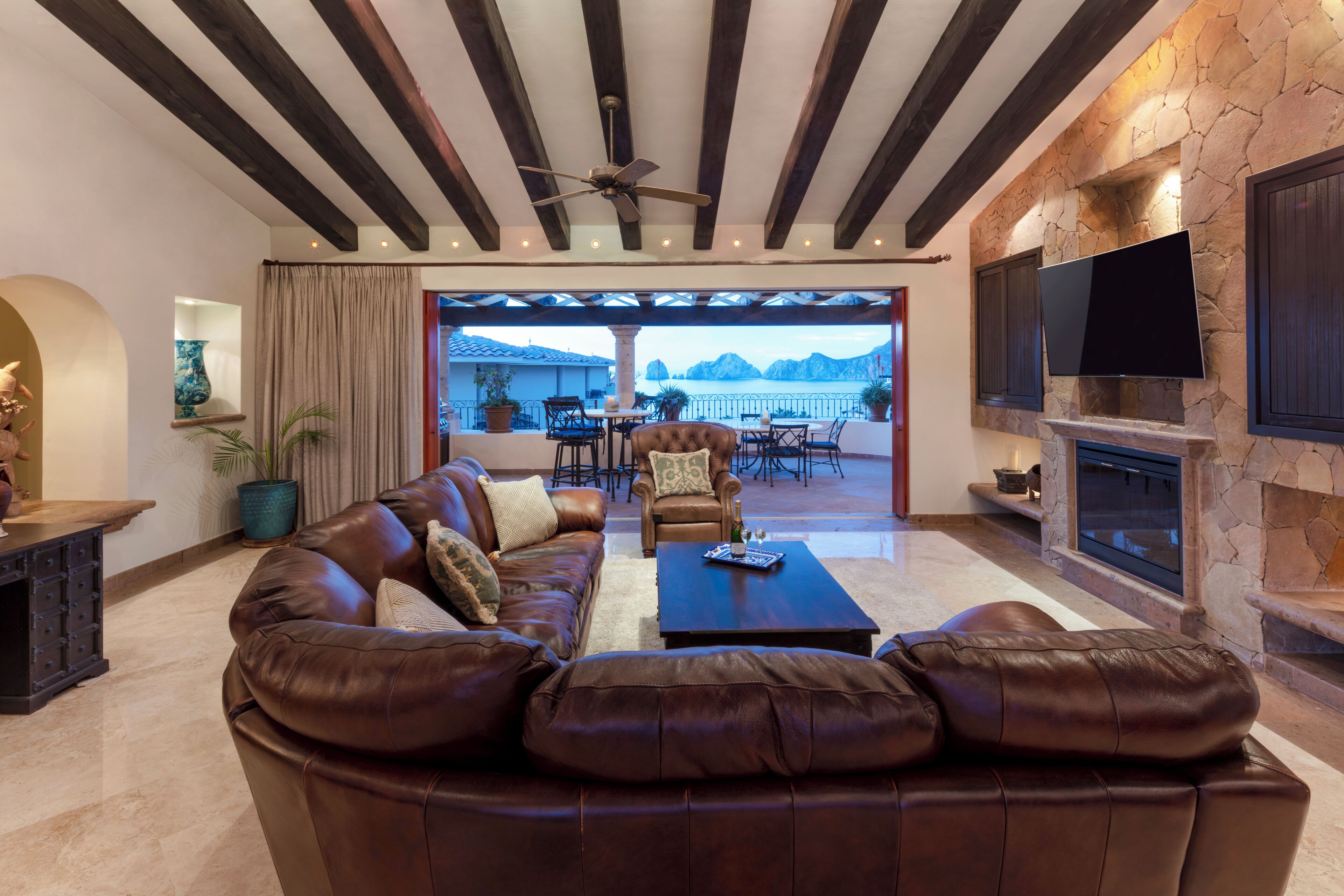 Un salón espacioso con sofá de cuero, una mesa de centro y una chimenea de piedra. Hay un televisor montado en la pared y un ventilador de techo.